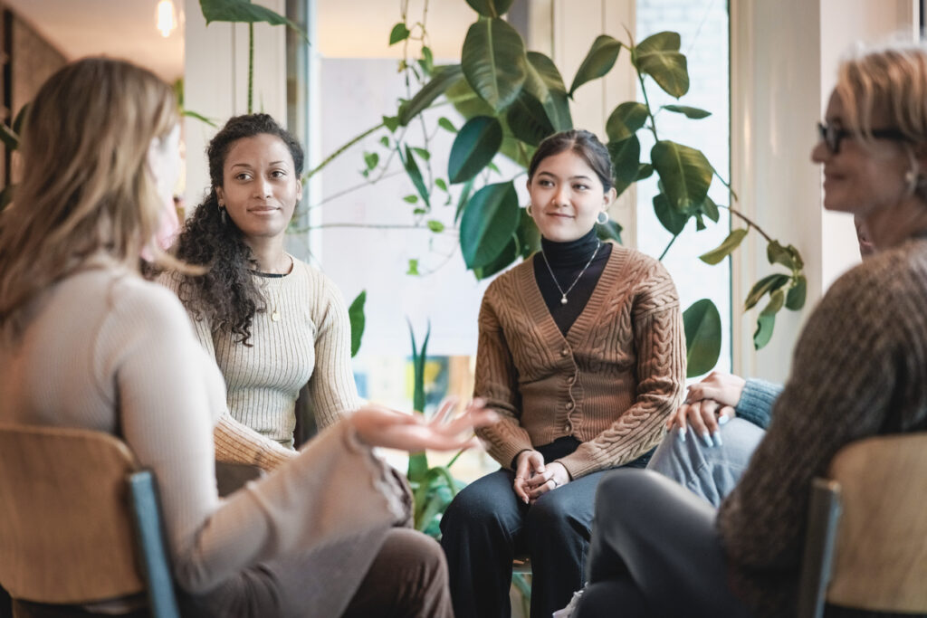 Woman sharing her experiences during a mental health group therapy meeting. Multiracial women participate in support group session sitting in a circle.