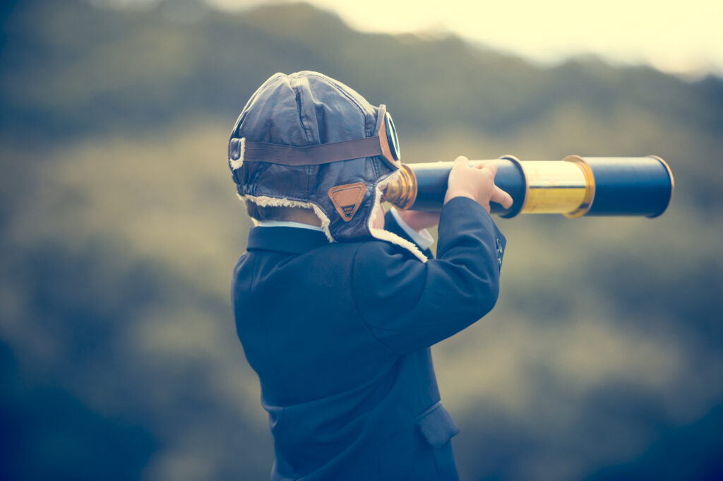 Young boy in a business suit with telescope. Small child wearing a full suit and holding a telescope. He is holding the telescope up to his eye with an aviator cap on. Business forecasting, innovation, leadership and planning concept. Shot outdoors with trees and grass in the background